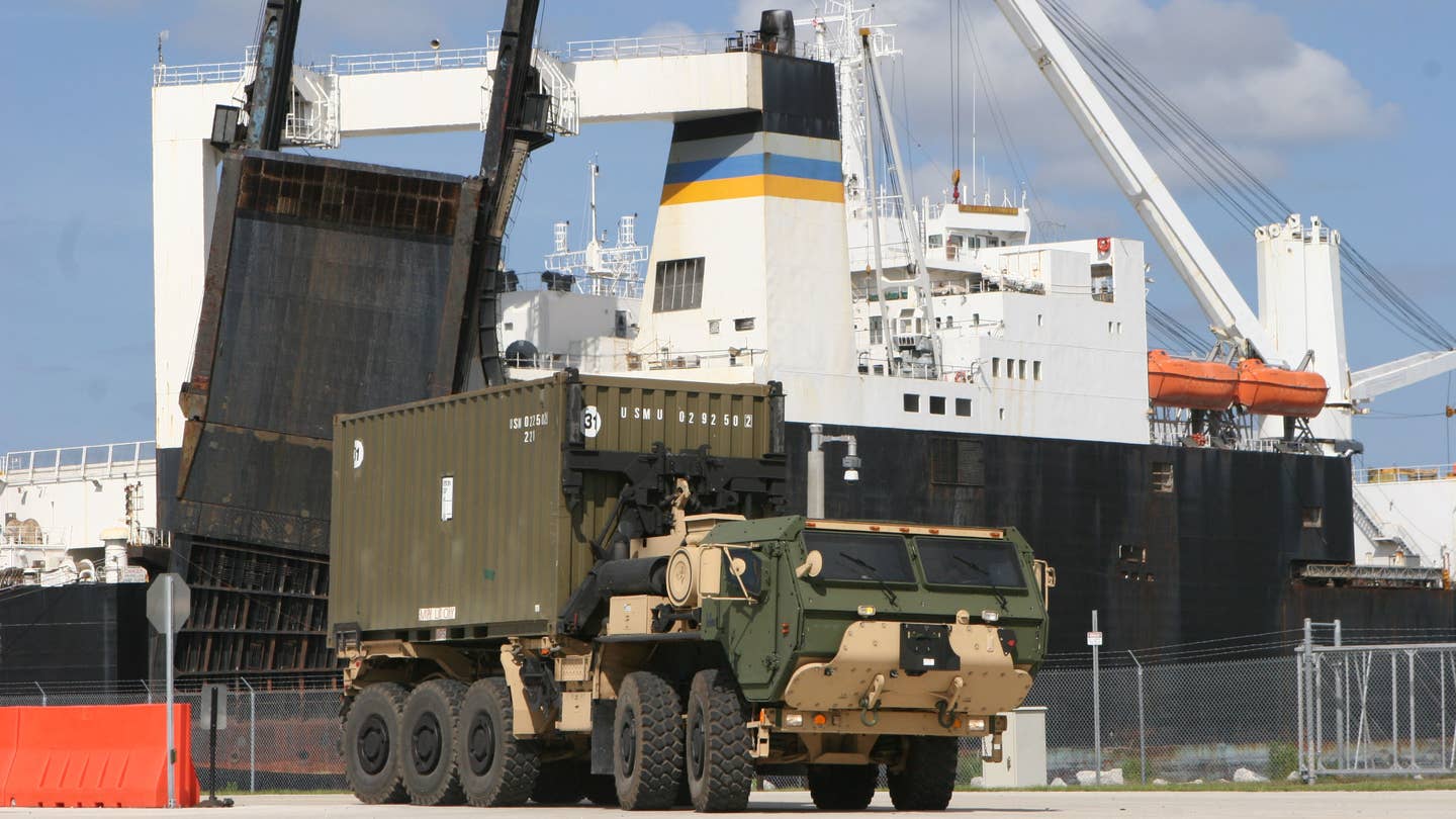 A US Marine Corps Logistics Vehicle System Replacement truck carrying a standard shipping container with a Navy logistics vessel in the background. The Navy is now working on a project to develop a containerized electronic warfare and electronic intelligence system that will work on various naval, air, and ground platforms.