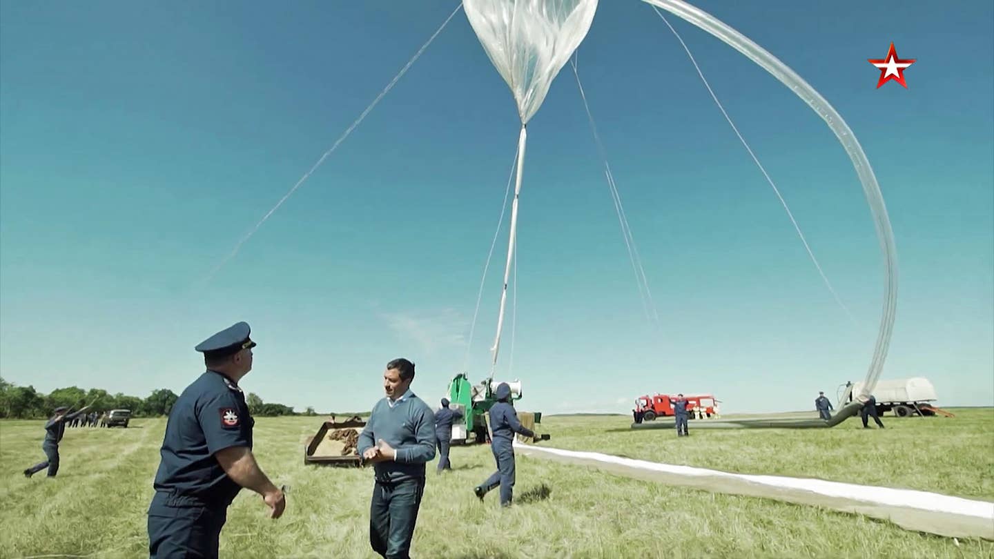 A partially inflated AN-S1 balloon begins to rise. The balloon continues to be filled through the large pipe on the right. The lower part of the envelope is held together with clamps, which it sheds as it continues to inflate. <em>TV Zvezda</em>