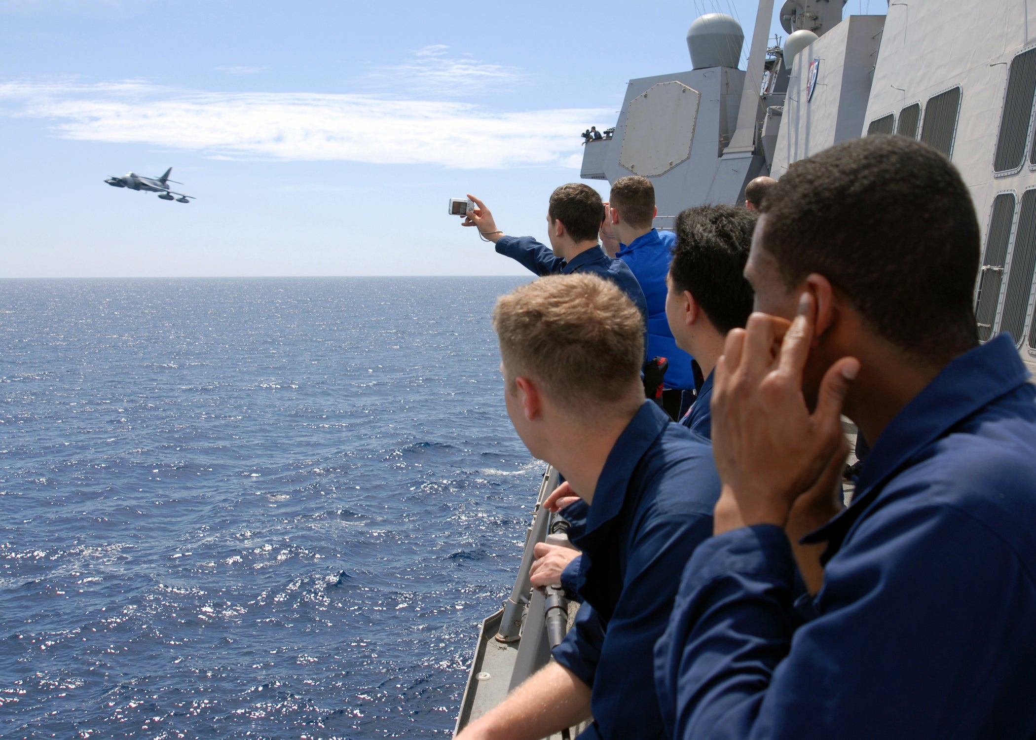 Sailors aboard the Arleigh Burke class guided missile destroyer USS Mustin gather around the rails to watch a Hawker Hunter training aircraft fly by the ship. Mustin is deployed with Destroyer Squadron 15.