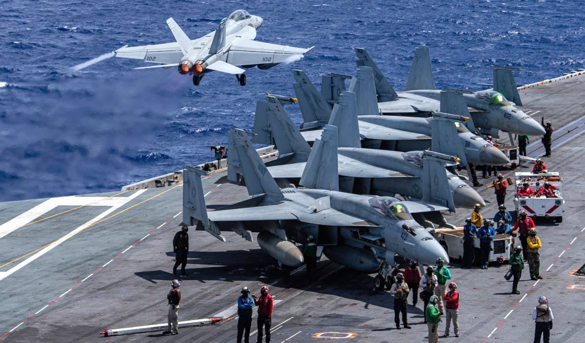 A row of U.S. Navy F/A-18Es are seen here on the deck of the supercarrier USS <em>Nimitz</em>. An F/A-18F is seen taking off in the background. <em>USN</em>