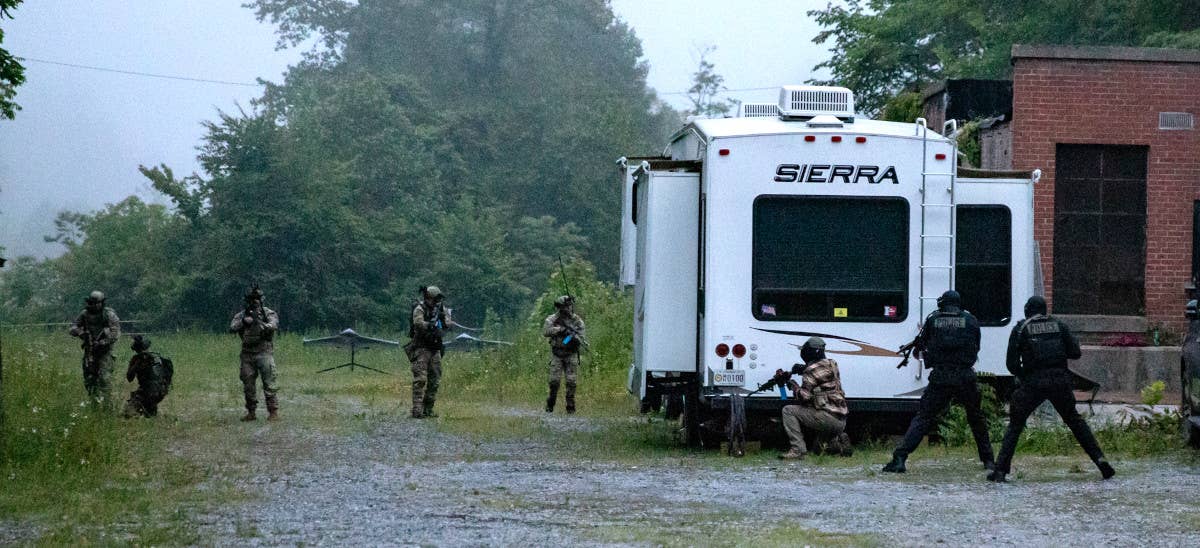 Personnel participating in Ridge Runner 23-01 advance through an area with members of the opposing force seen hiding behind a trailer. What appears to be two mock drones on stands are seen ion the background to the left. <em>Army National Guard </em>