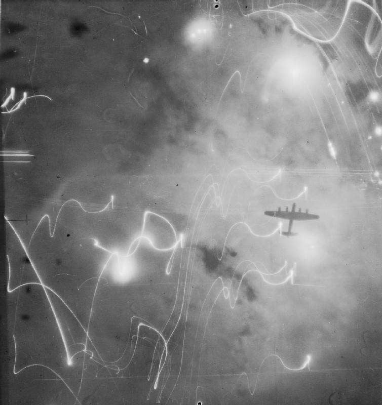 A well-known photo showing an RAF Bomber Command Avro Lancaster during an earlier attack on Hamburg, on the night of January 30/31, 1943. The bomber is silhouetted against flares, smoke, and explosions. This raid was the first time that H2S radar had been used by the Pathfinder aircraft to navigate the force to the target. <em>Crown Copyright</em>