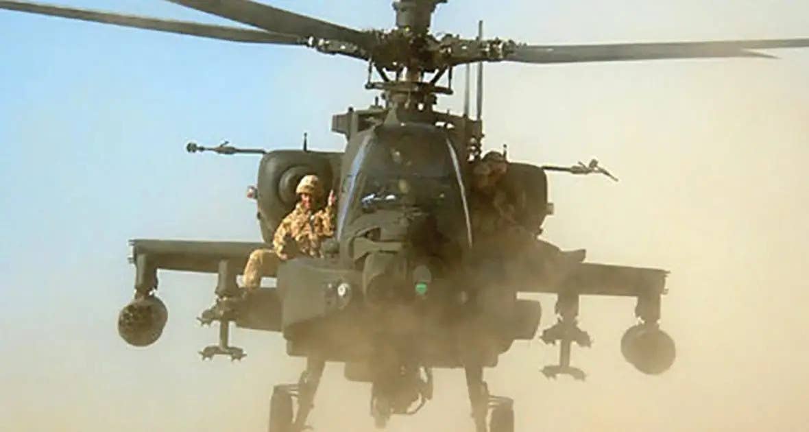 A British soldier rides strapped to one of the side sponsons on an AH-64 Apache attack helicopter. <em>Public Domain/UK MoD</em>