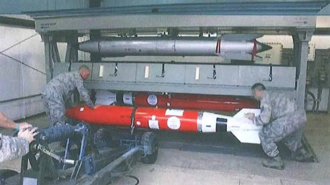 A picture showing a fit check of two B61-12 shapes, seen here with red-colored bodies, in a secure vault. A training version of an older B61 type is seen above. The distinctly different tail sections can be seen clearly here. <em>DOD via FOIA</em>