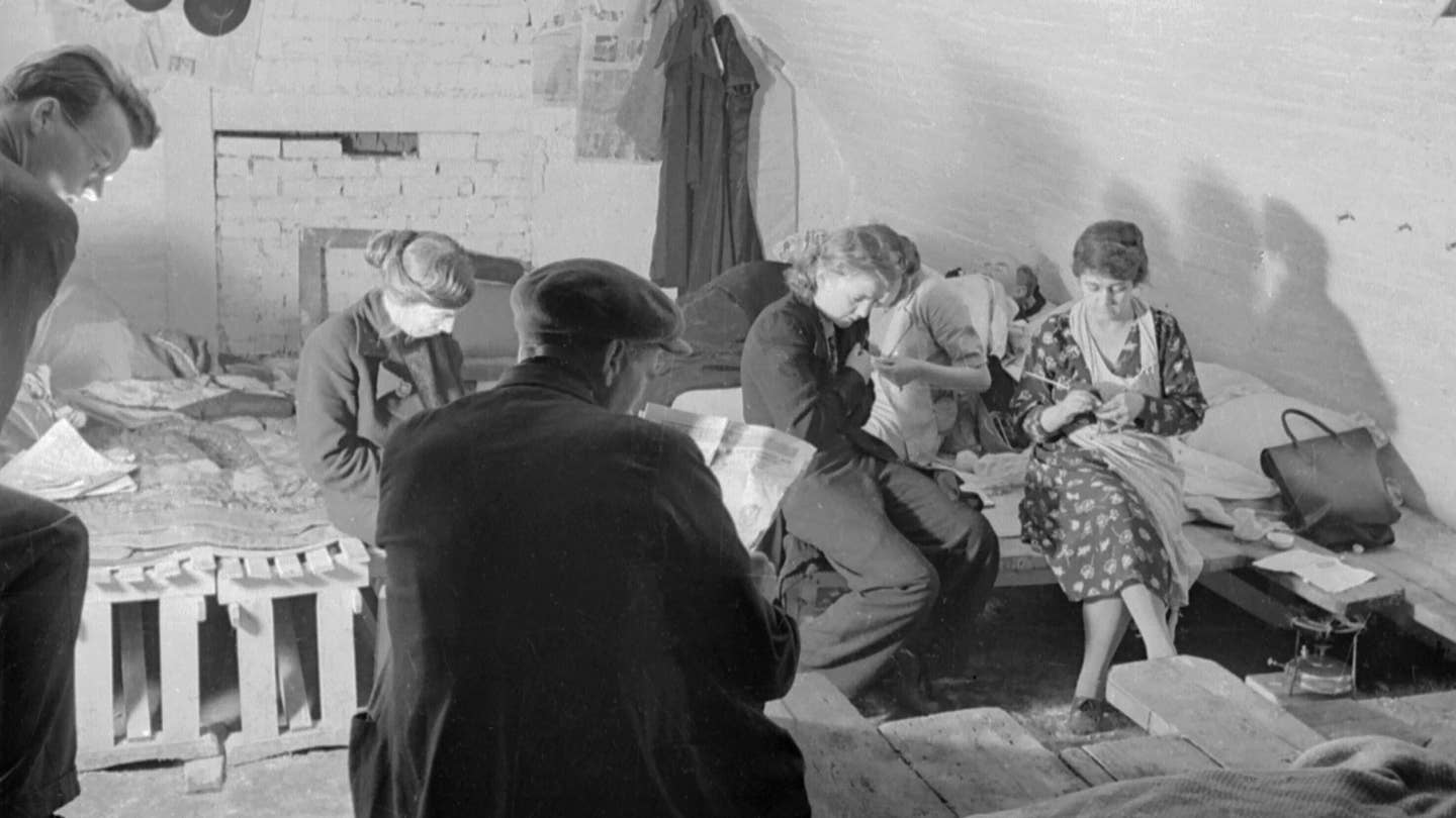 A small group of shelterers knit and read the evening newspaper in this small section of an air raid shelter under the railway arches, somewhere in South East London. Makeshift beds have been constructed from crates and planks of wood. This photograph was probably taken in November 1940