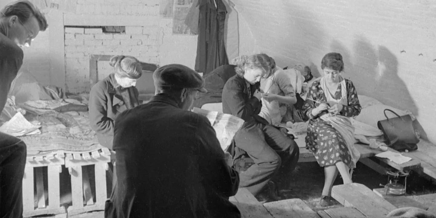 A small group of shelterers knit and read the evening newspaper in this small section of an air raid shelter under the railway arches, somewhere in South East London. Makeshift beds have been constructed from crates and planks of wood. This photograph was probably taken in November 1940
