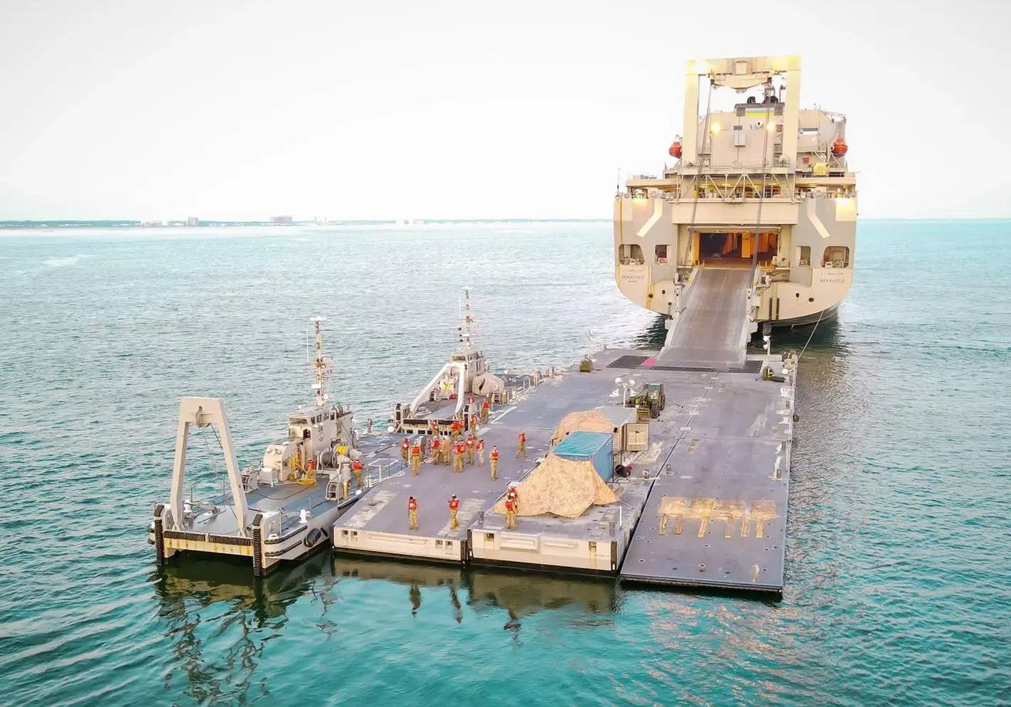 Elements of the Improved Navy Lighterage System (INLS) seen assembled during a training exercise. A roll-on/roll-off cargo ship is seen in the background with its stern ramp running right into the floating causeway. <em>USN</em>