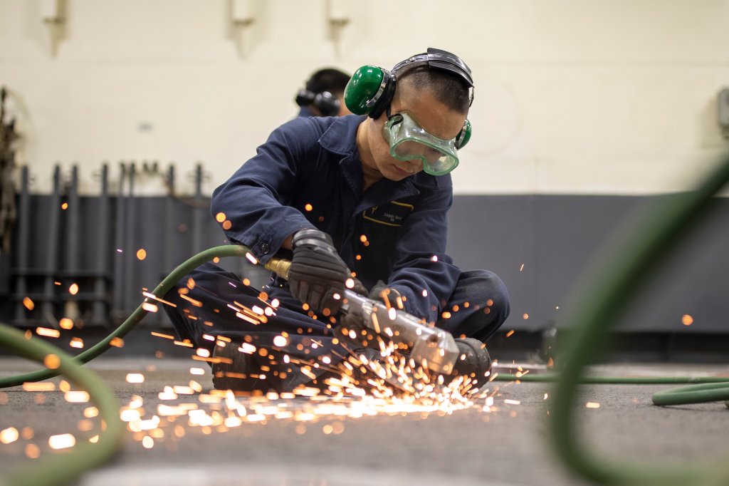 250313-N-KX492-1060 Seaman Dawie Guo, from Monterey Park, California, uses a grinder to remove rust from the deck in the vehicle stowage area aboard amphibious assault carrier USS Tripoli (LHA 7), Mar. 13, 2025. Tripoli is an America-class amphibious assault ship homeported in San Diego. (U.S. Navy photo by Mass Communication Specialist Seaman Paul LeClair)