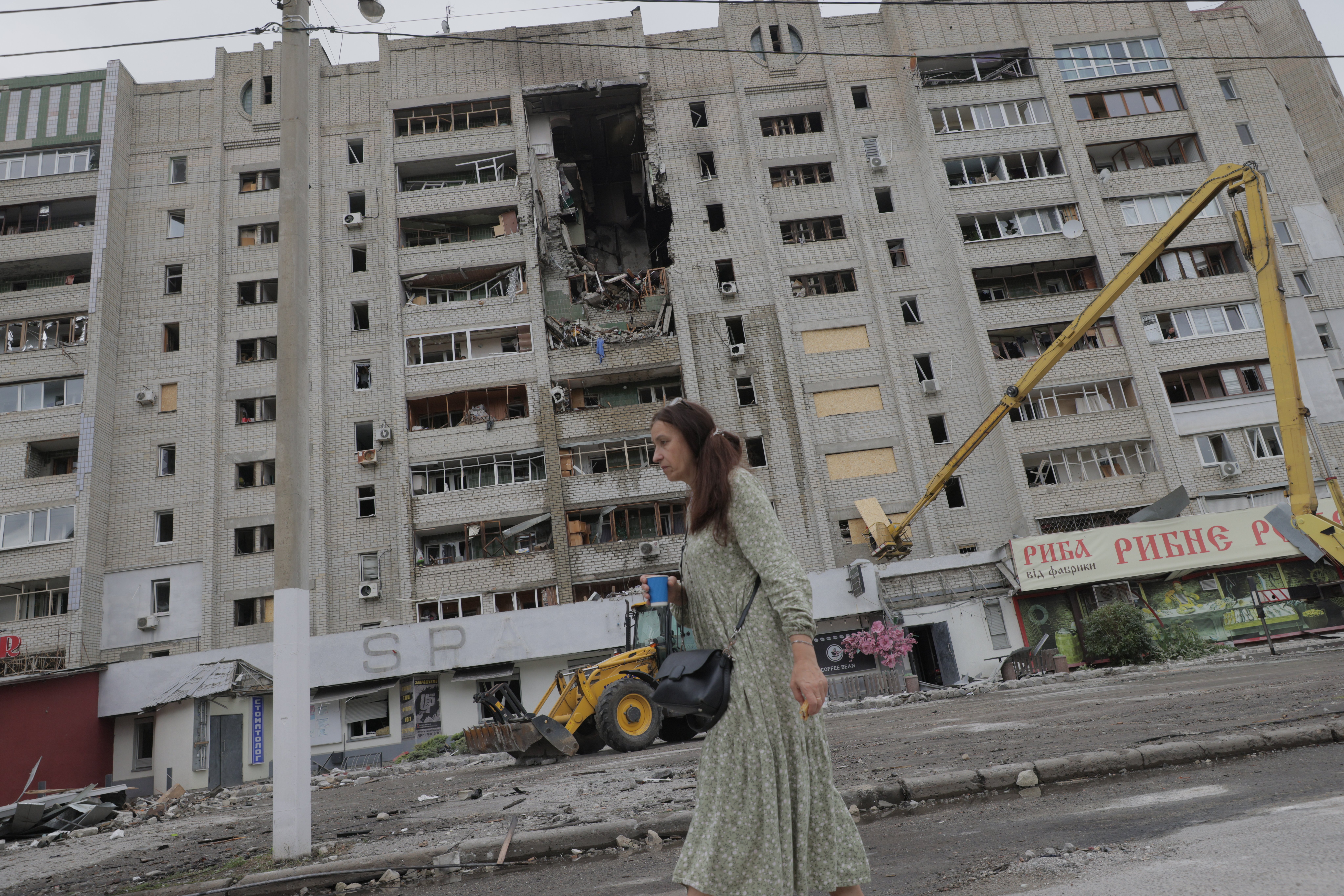 A woman walks past a severely damaged multi-story residential building in Kharkiv's Saltivka district, Ukraine, on August 9, 2026. The building sustained direct hits from drone strikes that destroyed multiple floors and killed two people, with dozens more injured; a yellow excavator sits among debris in the street below as cleanup and rescue efforts continue. (Photo by Yevhen Titov/NurPhoto via Getty Images)